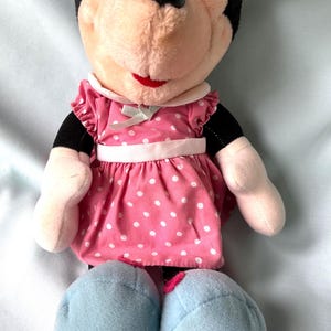 May include: A plush Minnie Mouse doll wearing a pink dress with white polka dots. The dress has a white collar and a light green bow. The doll has black ears, a red mouth, and light blue shoes.