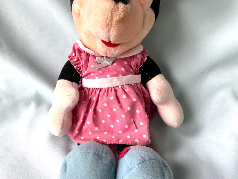 May include: A plush Minnie Mouse doll wearing a pink dress with white polka dots. The dress has a white collar and a light green bow. The doll has black ears, a red mouth, and light blue shoes.