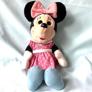 May include: A plush Minnie Mouse doll with black ears, a pink bow, and a pink polka-dot dress. The doll has light blue shoes and a smiling expression. The doll is a classic children's toy.