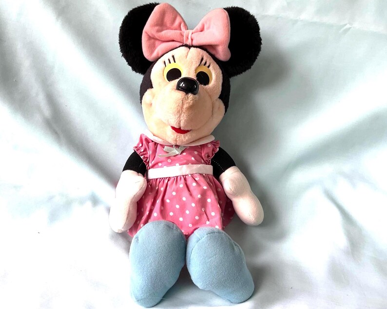 May include: A plush Minnie Mouse doll with black ears, a pink bow, and a pink polka-dot dress. The doll has light blue shoes and a smiling expression. The doll is a classic children's toy.