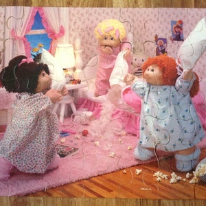 May include: A completed jigsaw puzzle featuring three Cabbage Patch Kids dolls in a bedroom setting. The dolls are in pajamas and holding pillows, with popcorn scattered on the pink carpet. The puzzle has a vintage aesthetic.