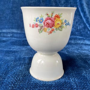 May include: White ceramic egg cup with a floral design featuring pink, yellow, and blue flowers. The egg cup has a rounded top and a pedestal base, perfect for serving eggs. The floral pattern adds a touch of elegance.