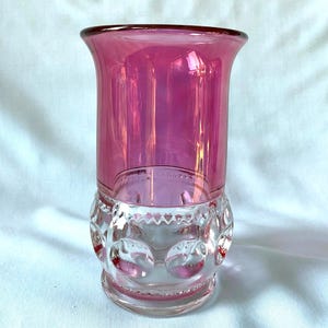 May include: A clear glass vase with a vibrant pink top and a decorative, patterned base. The vase has a flared rim and a cylindrical body. The base features a series of raised, circular designs. The vase is set against a white background.
