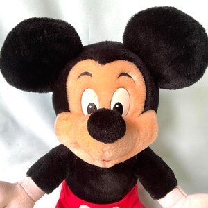 Puede incluir: Un peluche de Mickey Mouse con grandes orejas negras, cuerpo negro, pantalones cortos rojos y cara color canela. El juguete tiene ojos blancos, nariz negra y manos rosas. Un personaje cl&aacute;sico de Disney.