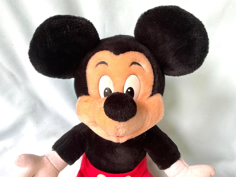 Puede incluir: Un peluche de Mickey Mouse con grandes orejas negras, cuerpo negro, pantalones cortos rojos y cara color canela. El juguete tiene ojos blancos, nariz negra y manos rosas. Un personaje cl&aacute;sico de Disney.