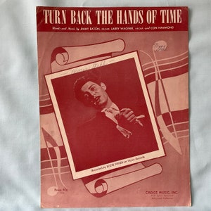 May include: Sheet music titled "Turn Back the Hands of Time" with a red and white color scheme. The cover features a photo of a man in a suit and tie. The text includes the names of the songwriters, the performer Eddie Fisher, and the publisher.