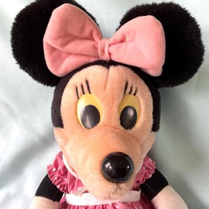 May include: A plush Minnie Mouse doll with black ears, a pink bow, and a pink and white polka-dot dress. The doll has large yellow eyes and a black nose. The doll is a classic Disney character.