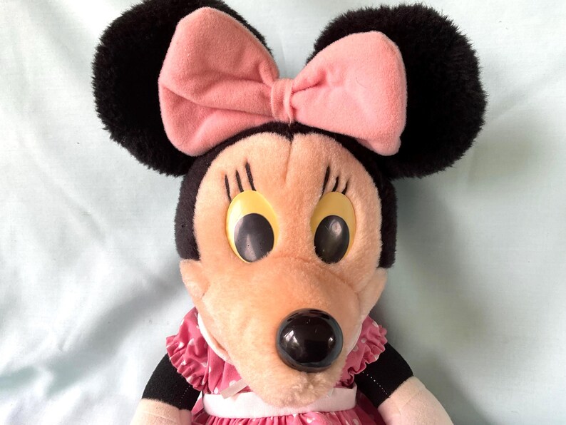 May include: A plush Minnie Mouse doll with black ears, a pink bow, and a pink and white polka-dot dress. The doll has large yellow eyes and a black nose. The doll is a classic Disney character.