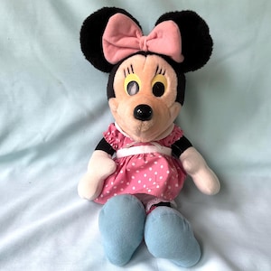 May include: A Minnie Mouse plush doll with black ears and a pink bow. She wears a pink dress with white polka dots, white gloves, and blue shoes. The doll has a black nose and yellow eyes.
