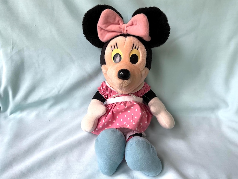 May include: A Minnie Mouse plush doll with black ears and a pink bow. She wears a pink dress with white polka dots, white gloves, and blue shoes. The doll has a black nose and yellow eyes.