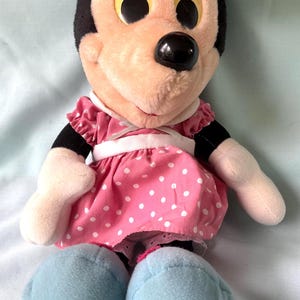 May include: A plush Minnie Mouse doll. The doll has a black head with large, round, yellow eyes and a black nose. She wears a pink dress with white polka dots and light blue shoes. The doll's arms and face are a light peach color.