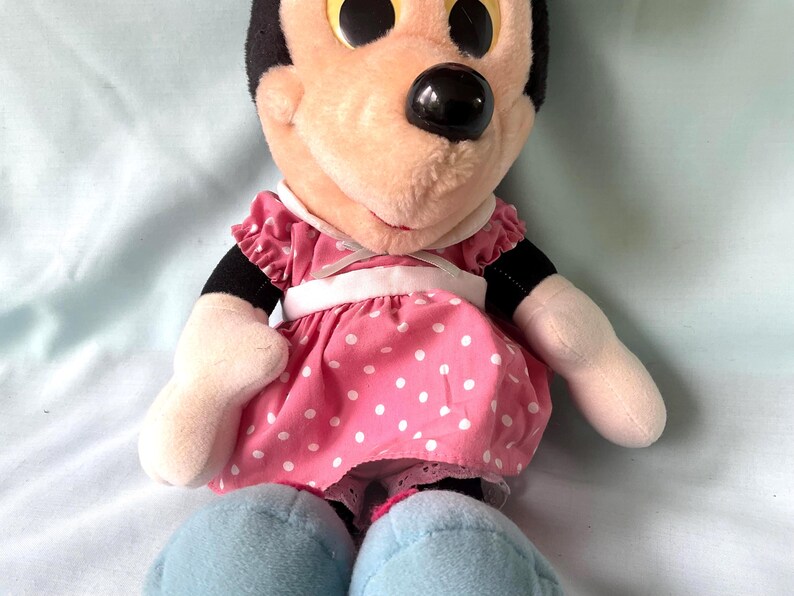 May include: A plush Minnie Mouse doll. The doll has a black head with large, round, yellow eyes and a black nose. She wears a pink dress with white polka dots and light blue shoes. The doll's arms and face are a light peach color.