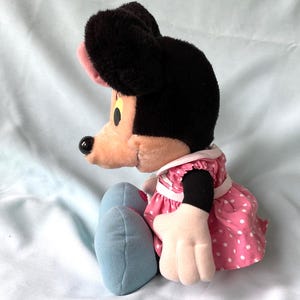 May include: A plush Minnie Mouse doll, featuring a black head and ears, a pink bow, and a pink and white polka dot dress. The doll has light blue shoes and white gloves. The doll is sitting on a light blue surface.