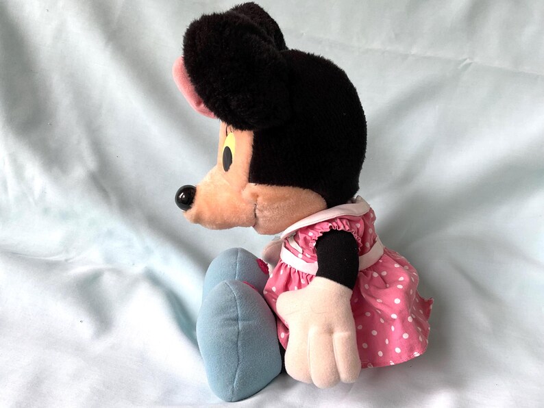 May include: A plush Minnie Mouse doll, featuring a black head and ears, a pink bow, and a pink and white polka dot dress. The doll has light blue shoes and white gloves. The doll is sitting on a light blue surface.