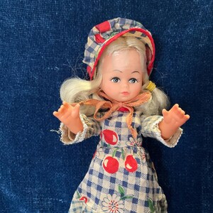 May include: A vintage doll with blonde hair, blue eyes, and a gingham bonnet. The doll wears a blue and white checkered dress with red cherry accents. The doll's arms are outstretched, and it has a peach-colored bow around its neck.