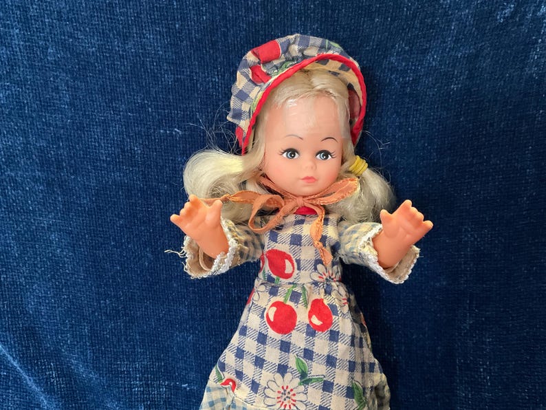 May include: A vintage doll with blonde hair, blue eyes, and a gingham bonnet. The doll wears a blue and white checkered dress with red cherry accents. The doll's arms are outstretched, and it has a peach-colored bow around its neck.