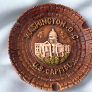 May include: A brown, circular ashtray featuring a relief of the U.S. Capitol building. The words "WASHINGTON D.C." and "U.S. CAPITOL" are embossed around the building. The ashtray has a textured, wood-like appearance.