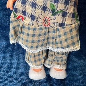 May include: A doll wearing a blue and tan checkered dress with a white lace trim. The dress features a floral design. The doll is also wearing matching checkered pants and white shoes.