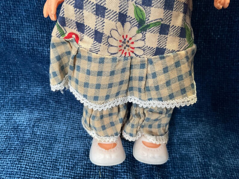 May include: A doll wearing a blue and tan checkered dress with a white lace trim. The dress features a floral design. The doll is also wearing matching checkered pants and white shoes.