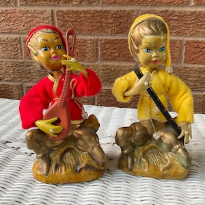 May include: Two vintage elf figurines. One elf wears a red outfit and holds a lute. The other elf wears a yellow outfit and holds a black instrument. Both elves have painted faces and are seated on a gold-colored base.
