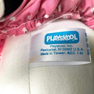 May include: Close-up of a Playskool plush toy. The toy has a white body and a pink polka-dot dress. A white tag with the Playskool logo and text is visible. The tag reads "Playskool, Inc. Pawtucket, RI 02862 U.S.A. Made in Taiwan, R.O.C. T-63."