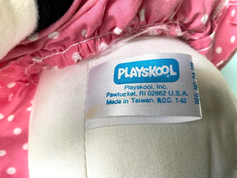 May include: Close-up of a Playskool plush toy. The toy has a white body and a pink polka-dot dress. A white tag with the Playskool logo and text is visible. The tag reads "Playskool, Inc. Pawtucket, RI 02862 U.S.A. Made in Taiwan, R.O.C. T-63."