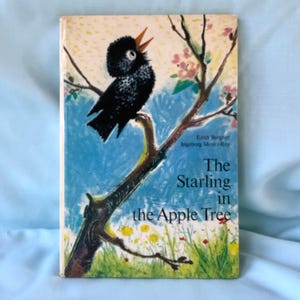 Puede incluir: Portada de libro vintage titulada "The Starling in the Apple Tree". La portada presenta un pájaro negro posado en una rama con flores rosas. El fondo es una mezcla de azul, amarillo y verde.