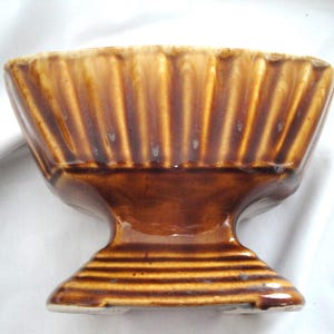 Puede incluir: Un cuenco de cerámica marrón con base de pedestal. El cuenco tiene un diseño acanalado y un acabado brillante. La base es de un tono marrón más oscuro con estrías horizontales. El cuenco es probablemente vintage o antiguo.