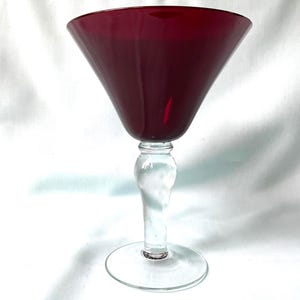 May include: A martini glass featuring a deep red, conical bowl set atop a clear, transparent glass stem and a circular base. The stem has a slightly bulbous, rounded shape in the center, providing a contrast to the dark red glass bowl.