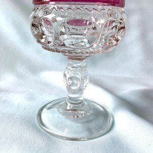 K&ouml;nnte beinhalten: Ein klares Glas-Goblet mit einem dekorativen Stiel und Fu&szlig;. Der Rand des Kelches ist tiefrosa. Die Schale des Kelches hat ein detailliertes Muster. Das Goblet wird wahrscheinlich zum Servieren von Getr&auml;nken verwendet.