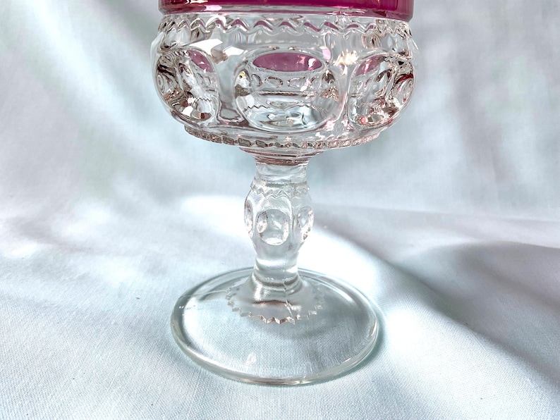 K&ouml;nnte beinhalten: Ein klares Glas-Goblet mit einem dekorativen Stiel und Fu&szlig;. Der Rand des Kelches ist tiefrosa. Die Schale des Kelches hat ein detailliertes Muster. Das Goblet wird wahrscheinlich zum Servieren von Getr&auml;nken verwendet.