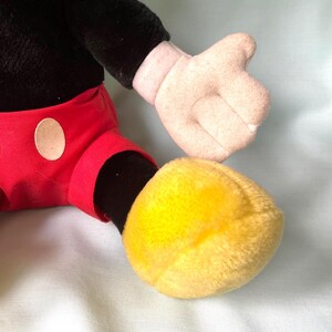 Puede incluir: Un peluche con la parte superior del cuerpo negra, pantalones cortos rojos con c&iacute;rculos blancos y pies amarillos. El juguete tiene una mano rosa claro y est&aacute; sobre un fondo azul claro. El juguete es un personaje cl&aacute;sico.