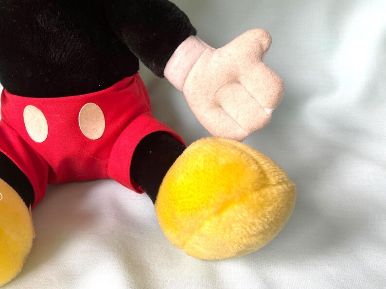 Puede incluir: Un peluche con la parte superior del cuerpo negra, pantalones cortos rojos con c&iacute;rculos blancos y pies amarillos. El juguete tiene una mano rosa claro y est&aacute; sobre un fondo azul claro. El juguete es un personaje cl&aacute;sico.