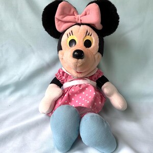 May include: A plush Minnie Mouse doll with black ears, a pink bow, and a pink dress with white polka dots. The doll has light blue shoes and white gloves. The doll is sitting on a light blue surface.
