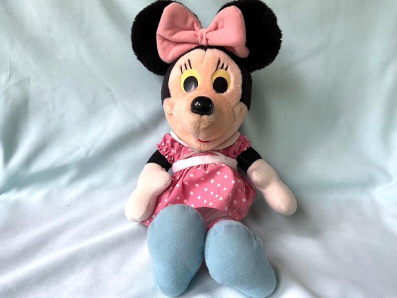 May include: A plush Minnie Mouse doll with black ears, a pink bow, and a pink dress with white polka dots. The doll has light blue shoes and white gloves. The doll is sitting on a light blue surface.
