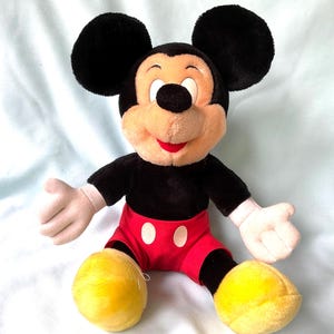 Puede incluir: Un peluche de Mickey Mouse. El juguete tiene grandes orejas negras, una cara bronceada y una boca roja. Viste una camisa negra, pantalones cortos rojos con botones blancos y zapatos amarillos. Los brazos y las manos son blancos.