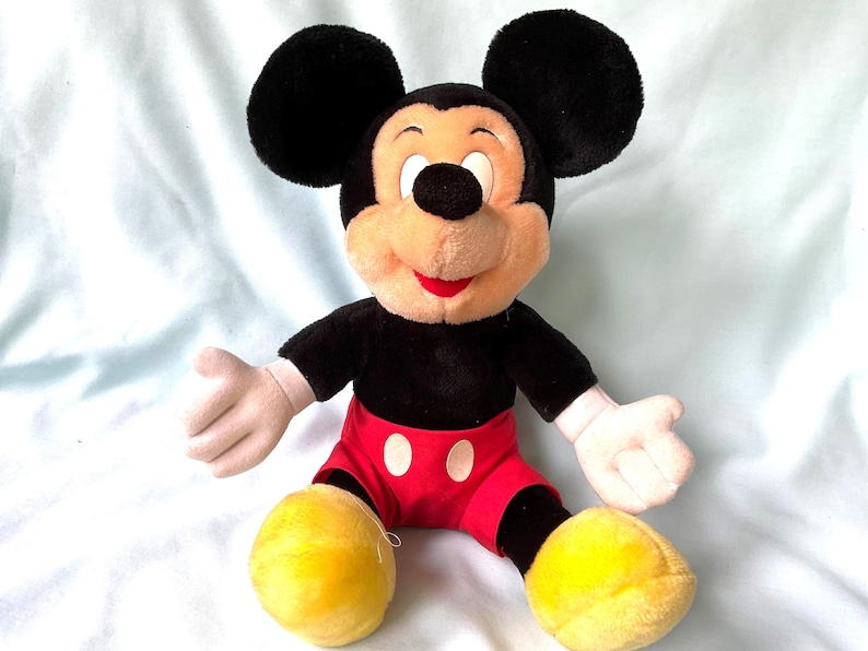 Puede incluir: Un peluche de Mickey Mouse. El juguete tiene grandes orejas negras, una cara bronceada y una boca roja. Viste una camisa negra, pantalones cortos rojos con botones blancos y zapatos amarillos. Los brazos y las manos son blancos.