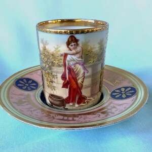 May include: A vintage teacup and saucer set. The cup and saucer are decorated with a classical scene featuring a woman in a red robe. The cup has a gold rim and the saucer has a pink and gold border with blue floral accents. The set is on a light blue surface.