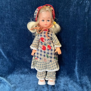 May include: A vintage doll with blonde hair and a red and white checkered bonnet. The doll wears a blue and white checkered dress with red cherry accents and matching pants. The doll is approximately 6 inches tall.