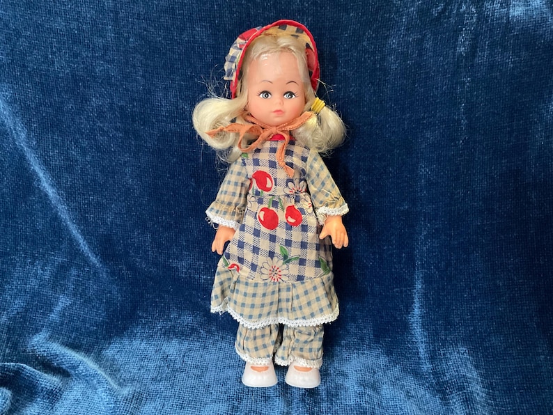 May include: A vintage doll with blonde hair and a red and white checkered bonnet. The doll wears a blue and white checkered dress with red cherry accents and matching pants. The doll is approximately 6 inches tall.