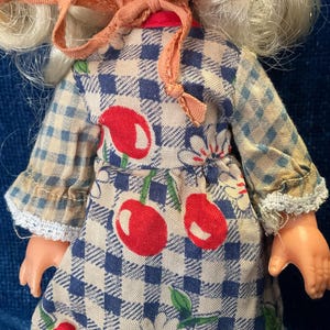 May include: A vintage doll wearing a blue and white checkered dress with red cherry and white flower accents. The doll has blonde hair and a pink bonnet. The dress has lace trim on the sleeves.