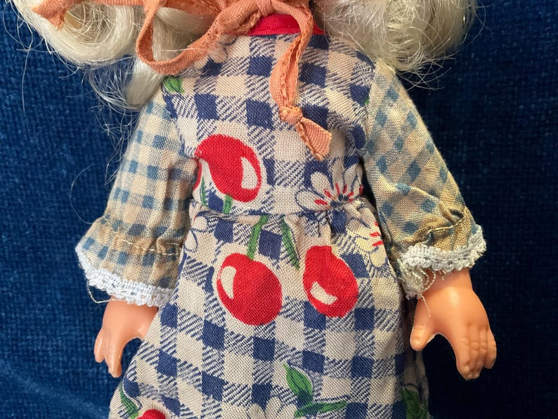 May include: A vintage doll wearing a blue and white checkered dress with red cherry and white flower accents. The doll has blonde hair and a pink bonnet. The dress has lace trim on the sleeves.