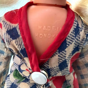 May include: Close-up of a vintage doll wearing a patterned kimono-style robe. The robe features a blue, red, and beige plaid design with a red trim. The doll's back is visible, with the text "MADE IN HONG-K" embossed on the plastic.