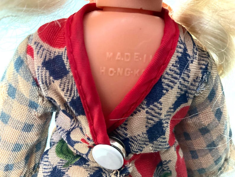 May include: Close-up of a vintage doll wearing a patterned kimono-style robe. The robe features a blue, red, and beige plaid design with a red trim. The doll's back is visible, with the text "MADE IN HONG-K" embossed on the plastic.