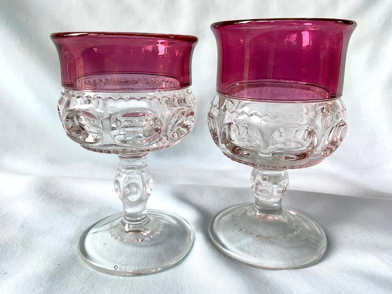K&ouml;nnte beinhalten: Zwei antike Weingl&auml;ser mit klaren, gemusterten Kelchen und leuchtend magentafarbenen R&auml;ndern. Die Stiele sind transparent mit dekorativen Details, und die B&ouml;den sind rund. Die Gl&auml;ser sind wahrscheinlich aus Glas und vor wei&szlig;em Hintergrund platziert.