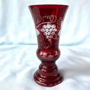 Op de afbeelding: Een robijnrode glazen vaas met een uitlopende bovenkant en een voetstuk. De vaas is versierd met geëtste witte druiven- en wijnrankenontwerpen. De vaas is ongeveer 18 cm hoog.