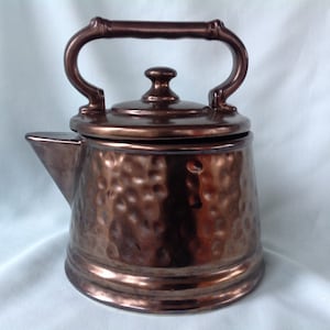 May include: A copper-colored ceramic teapot with a textured, hammered surface. It features a curved handle, a spout, and a lid with a small knob. The teapot has a classic design, suitable for serving tea.