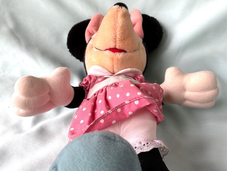May include: A plush Minnie Mouse doll with a smiling expression. She wears a pink dress with white polka dots, a white collar, and a matching bow. Her black ears and pink bow are visible.