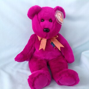 Puede incluir: Un oso de peluche magenta con nariz y ojos negros. Tiene una cinta color melocotón atada al cuello y una etiqueta en la oreja. El oso está sentado sobre una superficie azul claro.