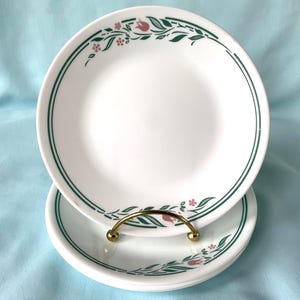 Könnte beinhalten: Zwei weiße Keramikteller mit einem grünen und rosa Blumendesign. Die Teller sind übereinander gestapelt und werden von einem goldfarbenen Metallständer gehalten. Die Teller haben einen grünen Rand und ein Blumenmuster aus rosa Tulpen und grünen Blättern.